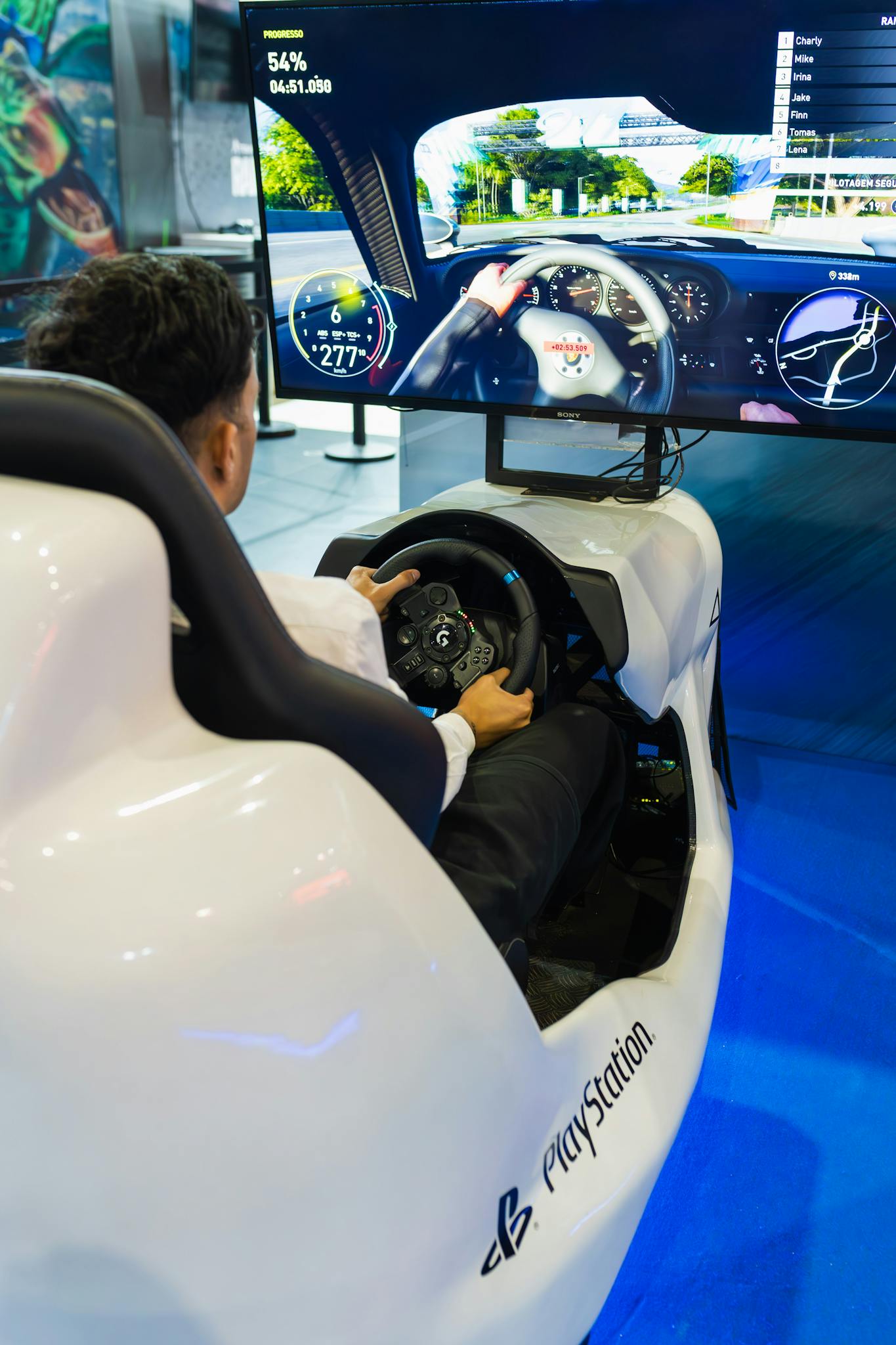 Man using PlayStation racing simulator at technology exhibition, showcasing gaming technology.